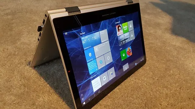 A convertible laptop in tent mode sits on a flat surface, displaying the Start screen with app tiles. The device has a metallic finish and sturdy hinges that allow the screen to fold back into the tent position, highlighting its 2‑in‑1 touchscreen design.