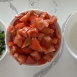 Ingredients for tomato jam: a plate of chopped spring onions (with sage, rosemary, minced ginger and garlic), a bowl of chopped tomatoes, a bowl of sugar on a white-veined kitchen counter