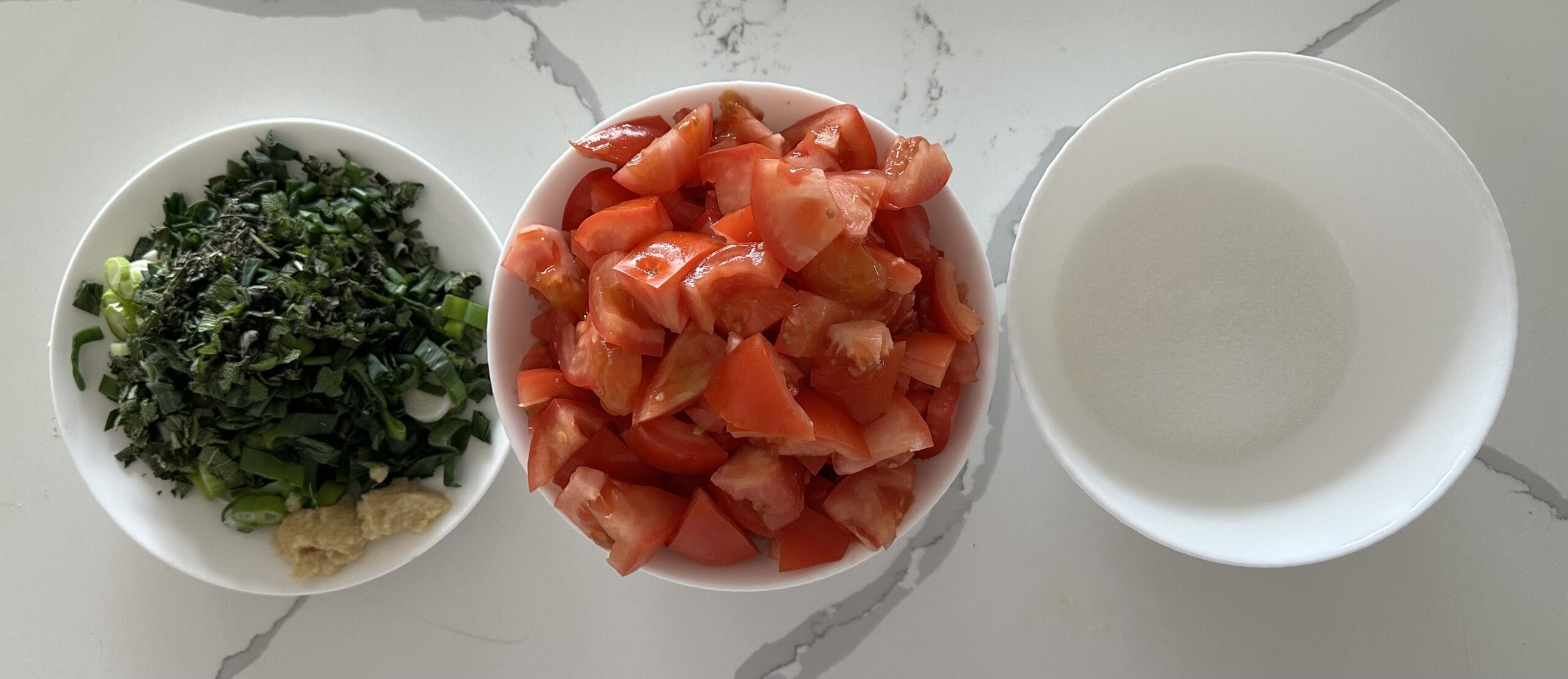 Ingredients for tomato jam: a plate of chopped spring onions (with sage, rosemary, minced ginger and garlic), a bowl of chopped tomatoes, a bowl of sugar on a white-veined kitchen counter