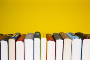 A row of hardcover books standing upright with their tops visible against a bright yellow background. The book covers vary in color, including blue, red, gray, brown, and black, creating a visually striking contrast with the yellow backdrop. The spines face away from the viewer, and while some text is partially visible, most titles are obscured. The composition highlights the diversity and tactile appeal of printed books in a modern, minimalist setting.