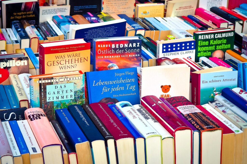 Stacks and rows of books, some standing on opening edge with spines showing, some standing on bottom edge with front cover showing.