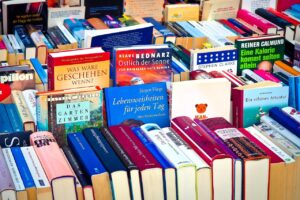 A vibrant and crowded book display at a fair or bookstore, featuring upright and stacked books in various languages, predominantly German. The covers are colorful and span genres including fiction, non-fiction, travel, and self-help. Visible titles include “Was wäre geschehen wenn?”, “Östlich der Sonne,” and “Lebensweisheiten für jeden Tag.” The eclectic mix and lively arrangement reflect a rich literary culture and diverse reading interests.