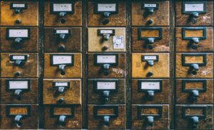 A vintage wooden library card catalog featuring dozens of small drawers arranged in a grid. Each drawer has a metal pull handle and a label holder displaying alphabetical ranges such as "WIE - WIK" and "ZUZ - ZYX." The wood is aged and worn, giving the catalog a rustic, historical feel. This type of furniture was traditionally used to organize index cards for books and resources in libraries before digital systems.