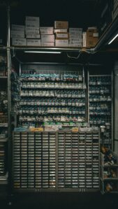 A meticulously organized section of a hardware or electronics store featuring a wall of hanging packages containing small components, likely for electronics or model building. Below the wall are two large cabinets with labeled drawers, some marked with Japanese and English terms like "TAMIYA" and "アーム 1号". Above the display are shelves stacked with cardboard boxes labeled with alphanumeric codes and Japanese text, indicating inventory. The overall setup suggests a specialized store catering to hobbyists or professionals.