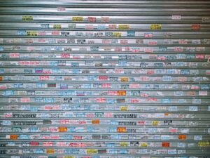 Metal roll‑up shutter door covered densely with colorful rectangular stickers displaying phone numbers and occasional Chinese characters, creating a chaotic patchwork of small service advertisements.