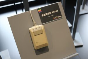 Beige Apple Macintosh M0100 mouse from 1984 displayed on a museum stand, with a single rectangular button and cord, accompanied by a label in Chinese identifying it as the Macintosh Mouse M0100