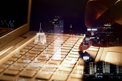 A close-up of a hand hovering above a laptop keyboard, blended with a nighttime cityscape of illuminated skyscrapers, creating a double‑exposure effect that suggests digital connectivity between technology and urban life.
