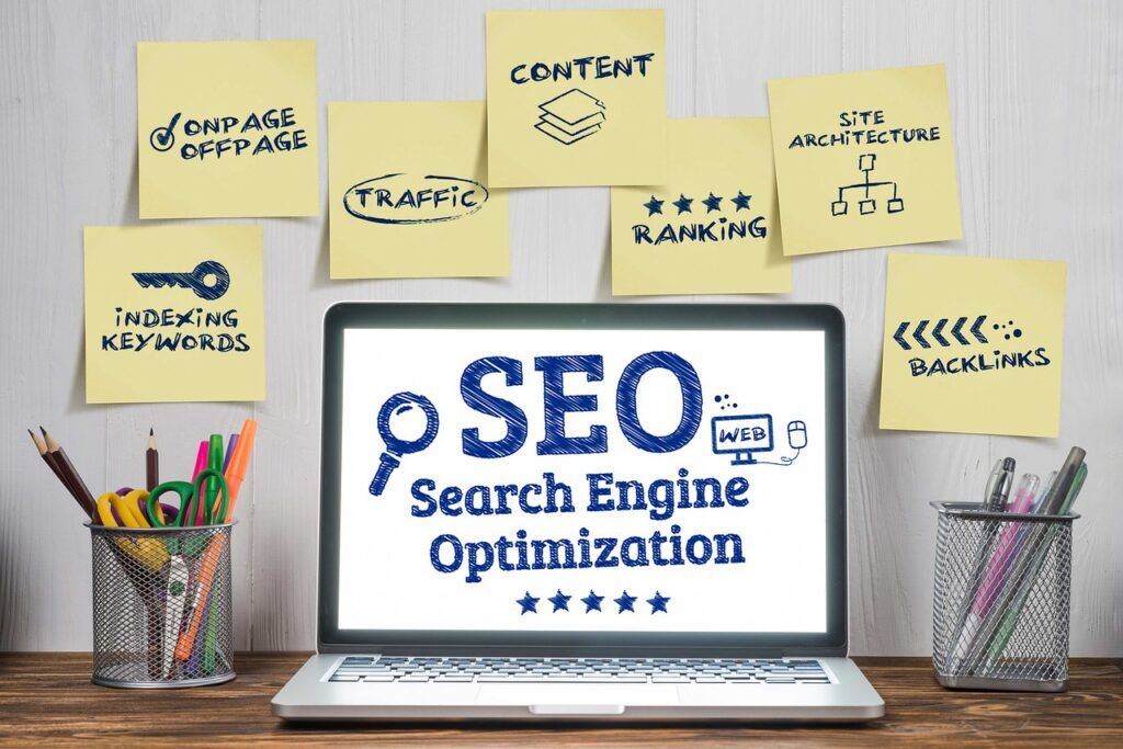 A laptop on a wooden desk displays the words “SEO Search Engine Optimization” with simple icons, surrounded by yellow sticky notes on the wall labeled with SEO concepts such as “ONPAGE OFFPAGE,” “TRAFFIC,” “CONTENT,” “SITE ARCHITECTURE,” “INDEXING KEYWORDS,” “RANKING,” and “BACKLINKS,” with pencil holders on either side of the laptop.