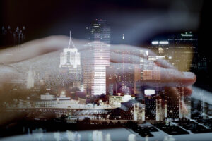 A close-up of a hand hovering above a laptop keyboard, blended with a nighttime cityscape of illuminated skyscrapers, creating a double‑exposure effect that suggests digital connectivity between technology and urban life.