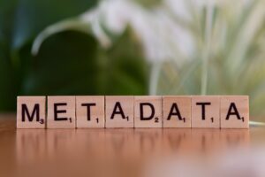 Wooden Scrabble-style tiles arranged in a row spell the word “METADATA,” each tile showing its letter and point value, set against a softly blurred green-and-white background.