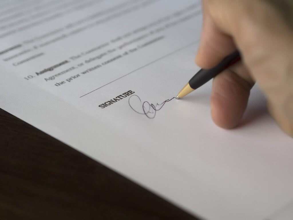 A close-up of a person signing a formal document with a black and gold pen. The paper shows printed text with numbered sections, and the word “SIGNATURE” is clearly visible above a handwritten signature being completed. The scene conveys the act of authorising or finalising an agreement.