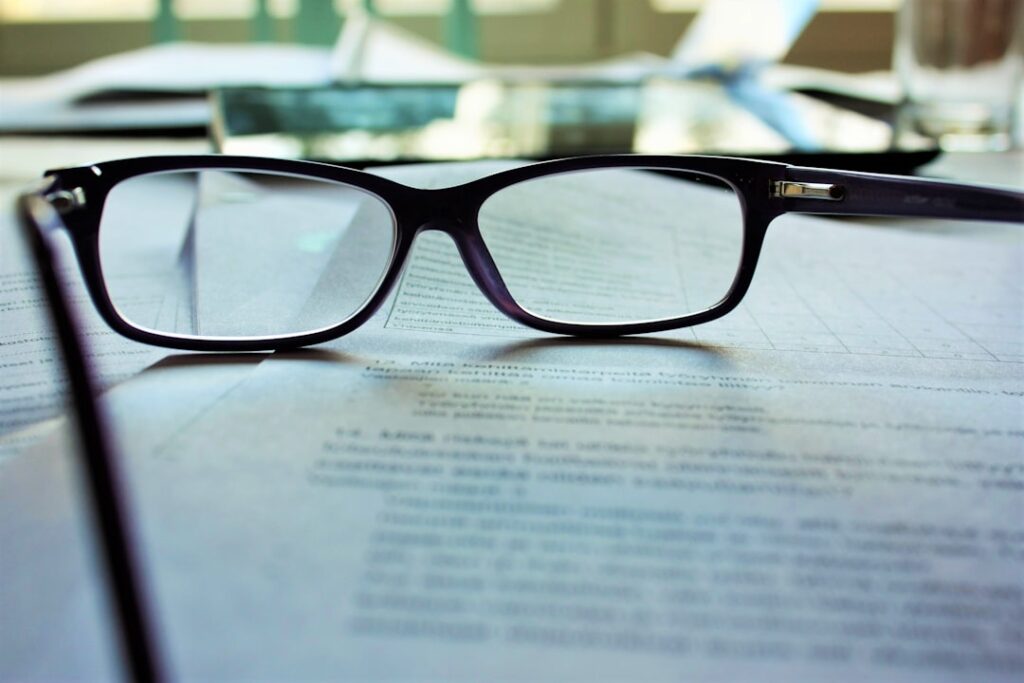 A close-up of black rectangular eyeglasses resting on printed documents. The lenses bring the text beneath them into sharp focus, while the surrounding words appear blurred, symbolising clarity and concentration. The background shows more papers and soft daylight, evoking a calm, studious workspace.