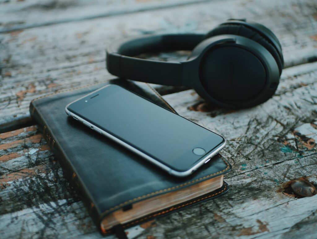 A close-up photo of a smartphone, a pair of black over-ear headphones, and a closed leather-bound book resting on a weathered wooden surface. The phone lies on top of the book, its reflective screen catching light, while the headphones sit slightly behind. The wood beneath shows peeling paint, cracks, and rusted bolts, giving the scene a rustic, textured look. The composition evokes a quiet moment of listening or reading, blending technology with timeless relaxation.