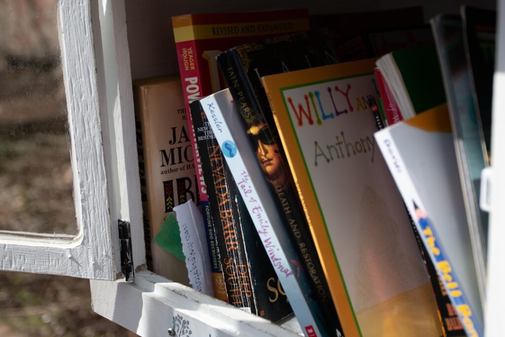 A close-up of a small white bookshelf or outdoor book cabinet filled with assorted books. The books are arranged vertically, their colorful spines creating a lively mix of hues. Sunlight filters through the glass door, suggesting a cozy, community book exchange or 'Little Free Library'.