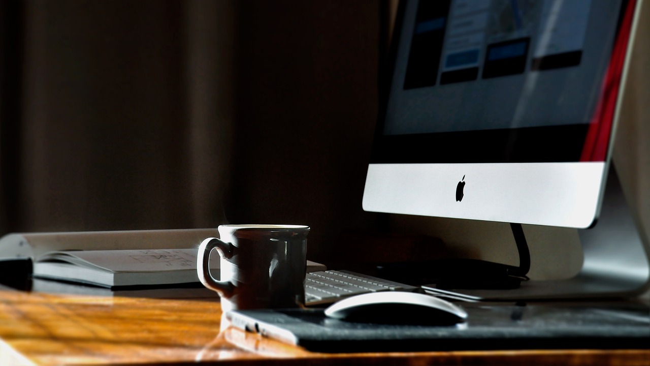 A tidy workspace featuring a computer on a wooden desk, accompanied by a keyboard, mouse, and a dark ceramic coffee mug. An open notebook lies nearby, and soft light from the side highlights the calm, focused atmosphere.