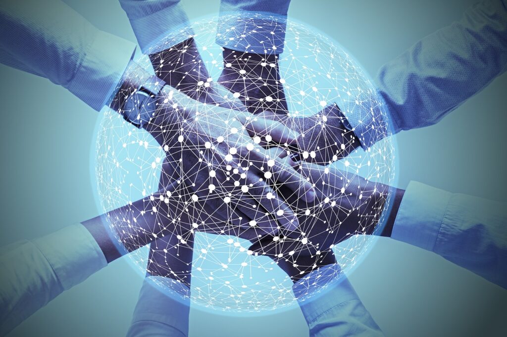 Several hands stacked together in a gesture of unity and teamwork, overlaid with a glowing digital network sphere made up of interconnected lines and nodes. The image symbolizes global collaboration, connectivity, and partnership in a technological or business context. The background features cool blue tones, enhancing the futuristic and cooperative theme.