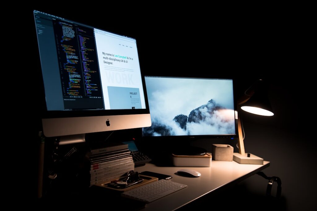 A modern workspace setup in a dark room featuring two computer monitors on a white desk. The left monitor shows colorful lines of code and a webpage introducing a UX/UI designer named Lee Campbell, while the right monitor displays a misty mountain scene. A desk lamp casts warm light over a keyboard, mouse, stacked books, and small accessories, creating a focused and minimalist atmosphere.