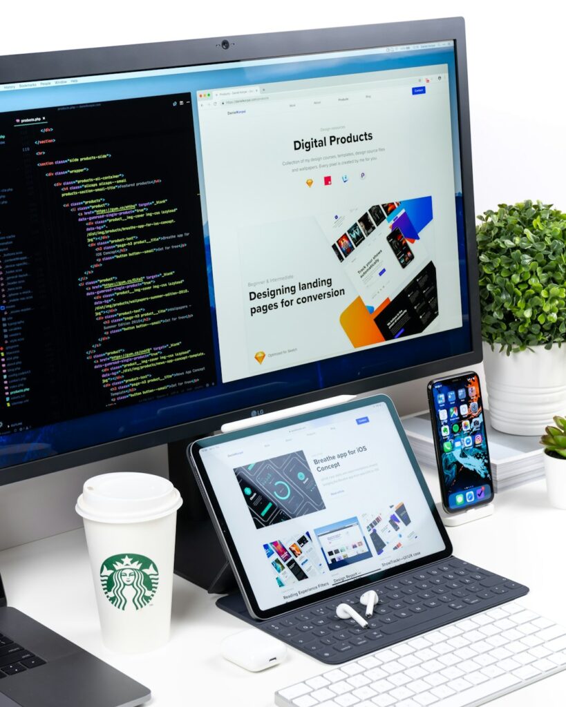 A modern workspace setup featuring a large monitor, tablet, smartphone, and laptop on a white desk. The monitor displays two windows: one showing HTML and CSS code, and the other a webpage titled “Digital Products” with text about designing landing pages for conversion. The tablet below shows another webpage titled “Breathe app for iOS Concept.” A Starbucks coffee cup, wireless earbuds, keyboard, and small green plants complete the scene. The image conveys a clean, professional environment for web development and digital design.
