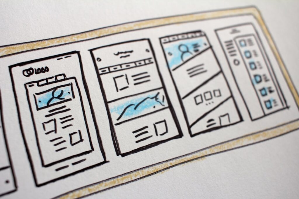 A close-up photo of a hand-drawn website wireframe sketch on paper. The drawing shows several rectangular layouts side by side, each representing different webpage designs with placeholders for logos, images, text blocks, and navigation bars. The sketches are outlined in black ink with touches of blue and yellow shading to highlight sections like banners and profile areas. The image conveys the concept of web design planning and user interface development.