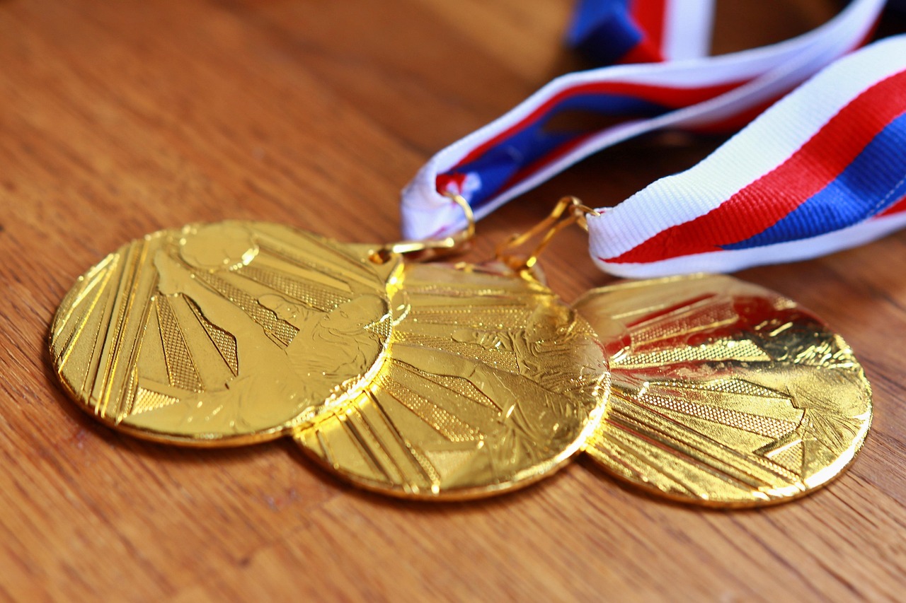 Three gold medals with embossed designs lie on a wooden surface. Each medal is attached to a red, white, and blue striped ribbon, symbolizing achievement and victory.